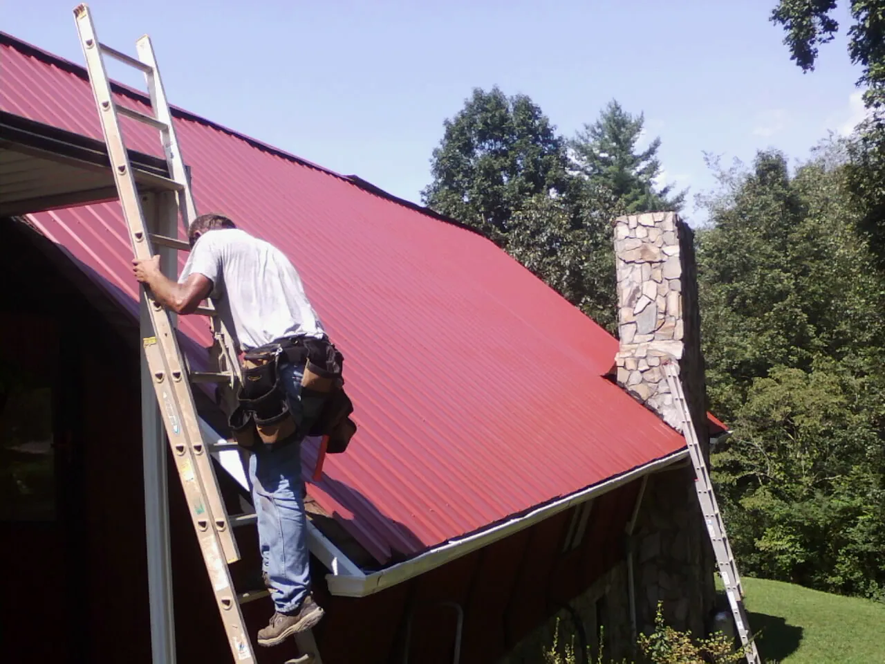 Roofer on a ladder beside a red metal roof and stone chimney — Soffit & Fascia Repair by Roofing Brothers in Glade Park